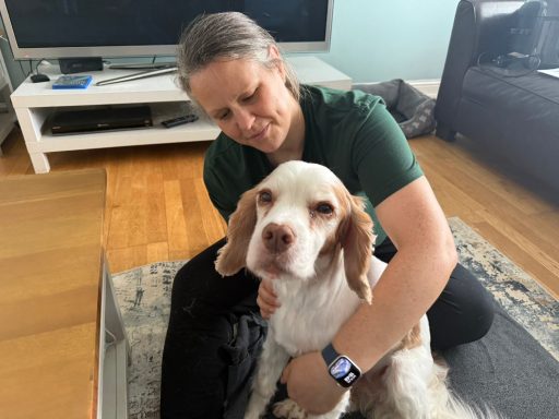 A woman sits on the floor, affectionately holding a dog in a living room setting.